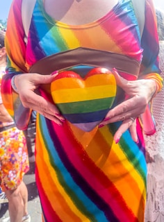 A person wearing a dress with rainbow stripes holds a heart-shaped object also featuring rainbow colors. The vibrant outfit and object are set against a background with other individuals. The person's fingernails are painted red, and a portion of a backpack strap is visible.