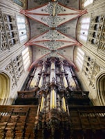 An intricately designed pipe organ is housed within a grand architectural setting. The ceiling above displays ornate patterns with floral motifs and pink arches. Tall, stained glass windows flank the organ, allowing light to filter through, enhancing the atmosphere with a sense of majesty.