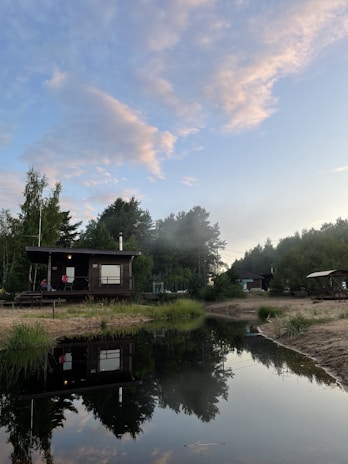 A wooden cabin is situated by a calm, reflective body of water, surrounded by lush greenery and tall trees. The sky is partly cloudy, with hues of blue and soft pinks. The scene is tranquil and picturesque, highlighted by the serene water reflection.