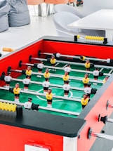 A foosball table with vibrant red sides, green playing surface, and two teams of miniature soccer players in yellow and white jerseys is positioned in a bright room. Surrounding the table are soft seating options, including bean bags and modern gray chairs, along with a white table.