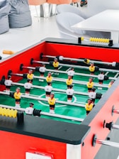 A foosball table with vibrant red sides, green playing surface, and two teams of miniature soccer players in yellow and white jerseys is positioned in a bright room. Surrounding the table are soft seating options, including bean bags and modern gray chairs, along with a white table.