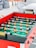 A foosball table with vibrant red sides, green playing surface, and two teams of miniature soccer players in yellow and white jerseys is positioned in a bright room. Surrounding the table are soft seating options, including bean bags and modern gray chairs, along with a white table.