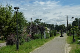 A paved pathway lined with tall lamp posts on both sides, surrounded by dense green vegetation and trees. In the distance, the path continues through a residential area with houses partially visible. Overhead, there are scattered clouds in a blue sky, and power lines stretch above the path.