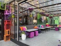 An outdoor patio area is furnished with modern sofas adorned with vibrant cushions in shades of orange, pink, and lime. Above, string lights hang across a clear ceiling, creating a welcoming atmosphere. The space is decorated with hanging planters filled with purple flowers. In the corner, colorful stacked crates add a playful touch.