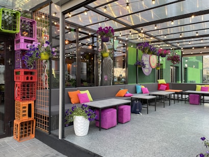 An outdoor patio area is furnished with modern sofas adorned with vibrant cushions in shades of orange, pink, and lime. Above, string lights hang across a clear ceiling, creating a welcoming atmosphere. The space is decorated with hanging planters filled with purple flowers. In the corner, colorful stacked crates add a playful touch.