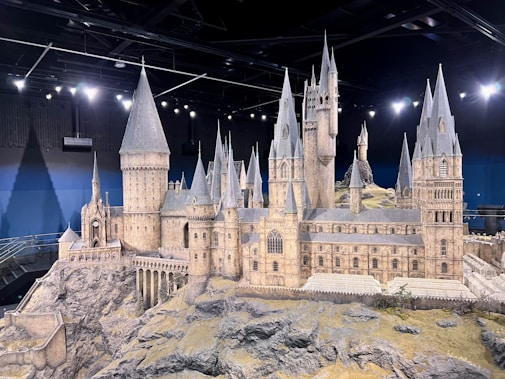 A detailed miniature model of a large, historic-style castle with multiple pointed towers and intricate architectural features. The structure is placed on a rocky terrain within an indoor setting, and bright lighting highlights the model's details.