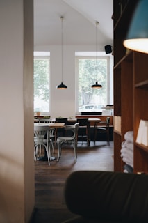 A cozy cafe interior with wooden furniture and minimalistic decor. The space features wooden tables and various styles of chairs, some with plants on them. Two large windows allow natural light to illuminate the space, while pendant lamps hang from the ceiling. Shelves with folded linens are visible on the right side.