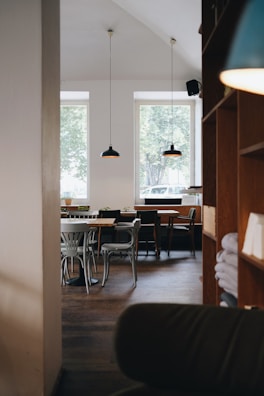 A cozy cafe interior with wooden furniture and minimalistic decor. The space features wooden tables and various styles of chairs, some with plants on them. Two large windows allow natural light to illuminate the space, while pendant lamps hang from the ceiling. Shelves with folded linens are visible on the right side.