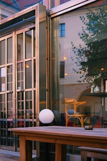 An outdoor wooden table and a round white lamp, situated in front of a large glass window reflecting the surrounding architecture. Inside, wooden chairs and a table with a small object on it are visible. The scene is framed by metal structures resembling an entrance or a facade.