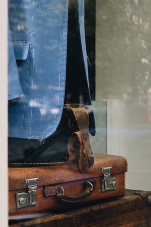 A tailored blue suit jacket draped over a vintage brown leather suitcase, visible through a glass display. The stitching on the jacket is evident, suggesting it is in a bespoke tailoring process. The reflections and lighting create a sophisticated and timeless feel.