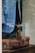 A tailored blue suit jacket draped over a vintage brown leather suitcase, visible through a glass display. The stitching on the jacket is evident, suggesting it is in a bespoke tailoring process. The reflections and lighting create a sophisticated and timeless feel.