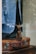 A tailored blue suit jacket draped over a vintage brown leather suitcase, visible through a glass display. The stitching on the jacket is evident, suggesting it is in a bespoke tailoring process. The reflections and lighting create a sophisticated and timeless feel.
