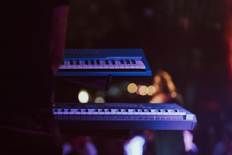 Dimly lit stage with two musicians surrounded by glowing synthesizers and keyboards.