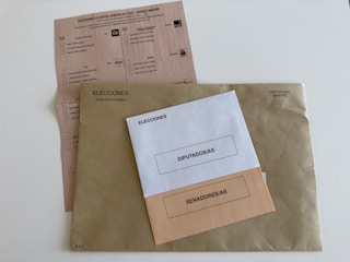 Three envelopes and a voting ballot are arranged on a flat surface. The largest envelope is brown with a text reading 'Elecciones Voto por Correo' and a stamp indicating it is urgent. On top of it, there are two smaller envelopes, one white labeled 'Diputados/as' and one beige labeled 'Senadores/as'. The voting ballot is partially visible beneath the envelopes, providing options for different political parties.