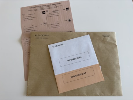 Three envelopes and a voting ballot are arranged on a flat surface. The largest envelope is brown with a text reading 'Elecciones Voto por Correo' and a stamp indicating it is urgent. On top of it, there are two smaller envelopes, one white labeled 'Diputados/as' and one beige labeled 'Senadores/as'. The voting ballot is partially visible beneath the envelopes, providing options for different political parties.
