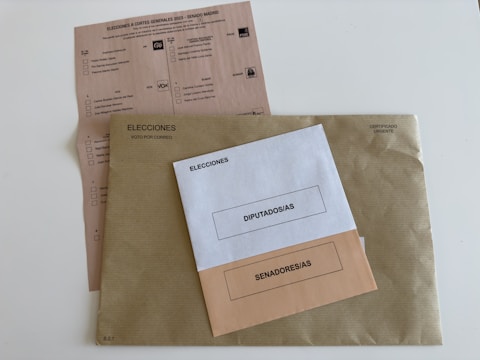 Three envelopes and a voting ballot are arranged on a flat surface. The largest envelope is brown with a text reading 'Elecciones Voto por Correo' and a stamp indicating it is urgent. On top of it, there are two smaller envelopes, one white labeled 'Diputados/as' and one beige labeled 'Senadores/as'. The voting ballot is partially visible beneath the envelopes, providing options for different political parties.
