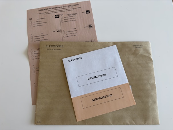 Three envelopes and a voting ballot are arranged on a flat surface. The largest envelope is brown with a text reading 'Elecciones Voto por Correo' and a stamp indicating it is urgent. On top of it, there are two smaller envelopes, one white labeled 'Diputados/as' and one beige labeled 'Senadores/as'. The voting ballot is partially visible beneath the envelopes, providing options for different political parties.