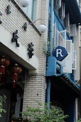 A street view of a building with a brick facade and blue accents. There are traditional Chinese decorations, including red lanterns with tassels. A sign with the letter 'R' is visible on the side of the building on a blue background. Plants and greenery are growing near the base.