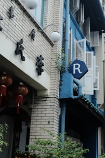 A street view of a building with a brick facade and blue accents. There are traditional Chinese decorations, including red lanterns with tassels. A sign with the letter 'R' is visible on the side of the building on a blue background. Plants and greenery are growing near the base.