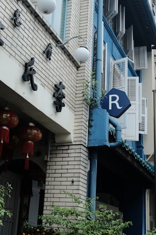 A street view of a building with a brick facade and blue accents. There are traditional Chinese decorations, including red lanterns with tassels. A sign with the letter 'R' is visible on the side of the building on a blue background. Plants and greenery are growing near the base.