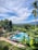 A serene landscape featuring a large, clear blue swimming pool surrounded by greenery, including tall palm trees and various other plants. The pool area is adorned with white umbrellas and lounge chairs, as well as a few stone statues. In the background, lush green hills and mountains are visible under a partly cloudy blue sky.