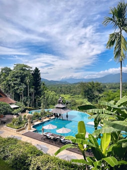 A serene landscape featuring a large, clear blue swimming pool surrounded by greenery, including tall palm trees and various other plants. The pool area is adorned with white umbrellas and lounge chairs, as well as a few stone statues. In the background, lush green hills and mountains are visible under a partly cloudy blue sky.