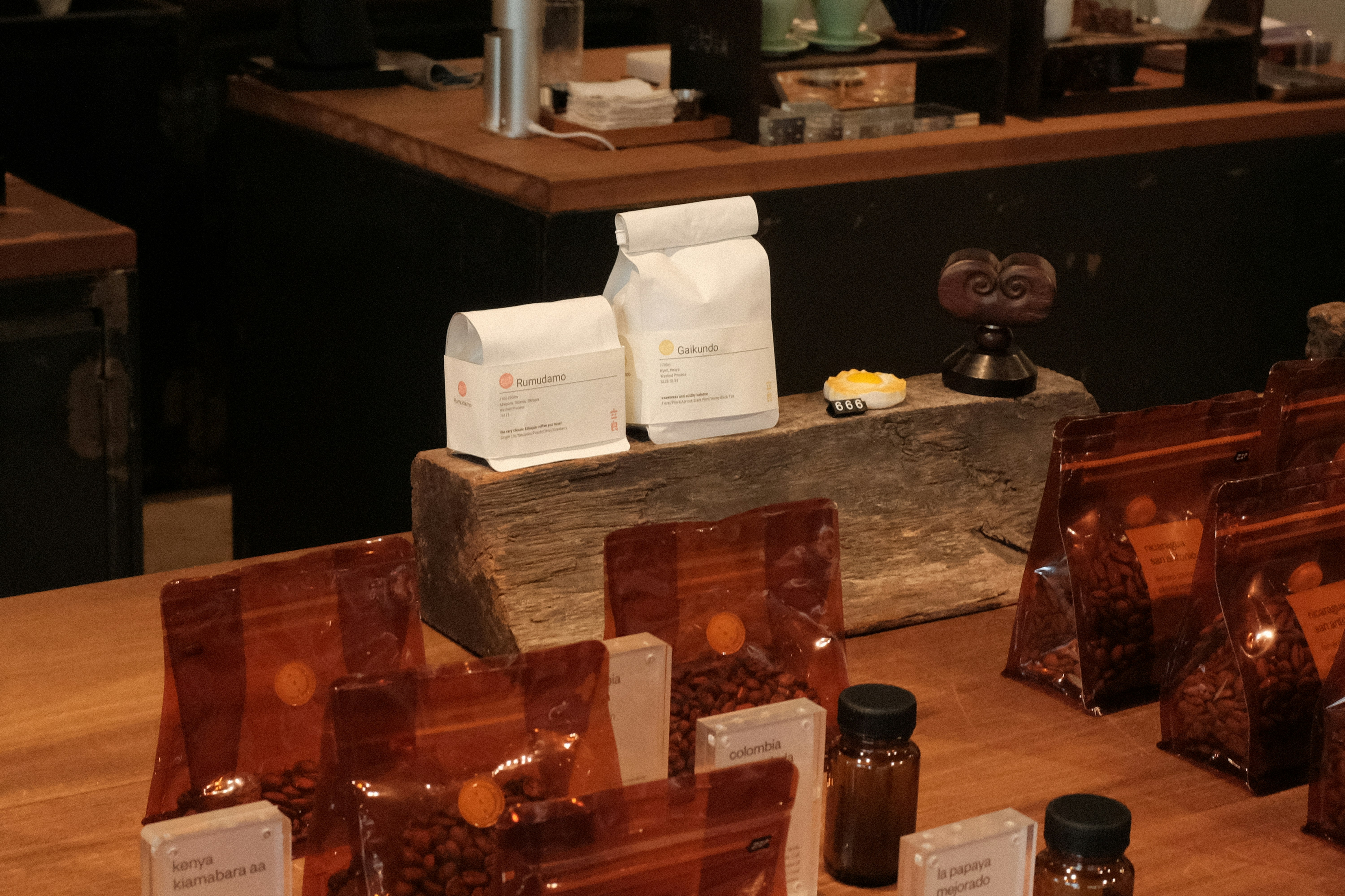 A variety of coffee products displayed on a wooden counter. Several sealed brown bags containing coffee beans are visible alongside two white bags labeled 'Rumudamo' and 'Gakuindo.' The setup includes a rustic piece of wood serving as a stand, and small brown and clear bottles are placed around the bags. The background includes shelves with cups, utensils, and additional coffee-related items.