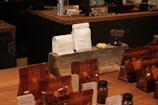 A variety of coffee products displayed on a wooden counter. Several sealed brown bags containing coffee beans are visible alongside two white bags labeled 'Rumudamo' and 'Gakuindo.' The setup includes a rustic piece of wood serving as a stand, and small brown and clear bottles are placed around the bags. The background includes shelves with cups, utensils, and additional coffee-related items.