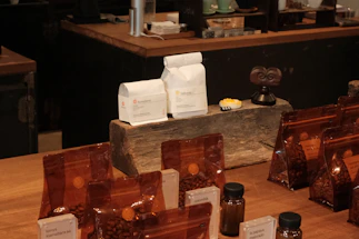 A variety of coffee products displayed on a wooden counter. Several sealed brown bags containing coffee beans are visible alongside two white bags labeled 'Rumudamo' and 'Gakuindo.' The setup includes a rustic piece of wood serving as a stand, and small brown and clear bottles are placed around the bags. The background includes shelves with cups, utensils, and additional coffee-related items.