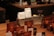 A variety of coffee products displayed on a wooden counter. Several sealed brown bags containing coffee beans are visible alongside two white bags labeled 'Rumudamo' and 'Gakuindo.' The setup includes a rustic piece of wood serving as a stand, and small brown and clear bottles are placed around the bags. The background includes shelves with cups, utensils, and additional coffee-related items.