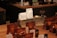 A variety of coffee products displayed on a wooden counter. Several sealed brown bags containing coffee beans are visible alongside two white bags labeled 'Rumudamo' and 'Gakuindo.' The setup includes a rustic piece of wood serving as a stand, and small brown and clear bottles are placed around the bags. The background includes shelves with cups, utensils, and additional coffee-related items.