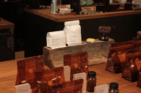 A variety of coffee products displayed on a wooden counter. Several sealed brown bags containing coffee beans are visible alongside two white bags labeled 'Rumudamo' and 'Gakuindo.' The setup includes a rustic piece of wood serving as a stand, and small brown and clear bottles are placed around the bags. The background includes shelves with cups, utensils, and additional coffee-related items.