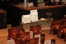 A variety of coffee products displayed on a wooden counter. Several sealed brown bags containing coffee beans are visible alongside two white bags labeled 'Rumudamo' and 'Gakuindo.' The setup includes a rustic piece of wood serving as a stand, and small brown and clear bottles are placed around the bags. The background includes shelves with cups, utensils, and additional coffee-related items.