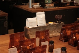 A variety of coffee products displayed on a wooden counter. Several sealed brown bags containing coffee beans are visible alongside two white bags labeled 'Rumudamo' and 'Gakuindo.' The setup includes a rustic piece of wood serving as a stand, and small brown and clear bottles are placed around the bags. The background includes shelves with cups, utensils, and additional coffee-related items.