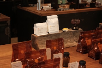 A variety of coffee products displayed on a wooden counter. Several sealed brown bags containing coffee beans are visible alongside two white bags labeled 'Rumudamo' and 'Gakuindo.' The setup includes a rustic piece of wood serving as a stand, and small brown and clear bottles are placed around the bags. The background includes shelves with cups, utensils, and additional coffee-related items.