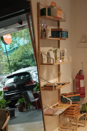 A wall-mounted wooden shelf is filled with various boxed items and packages. These shelves are part of an indoor space with a visible outdoor area through a nearby window or door. A black car is parked outside, and there's a fire extinguisher on the wall near the shelf. Some plants and chairs can be seen outside.