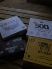 Four different board game boxes placed on a wooden surface. The designs feature colorful and graphic artwork. Each box appears to have distinct themes, represented by unique titles and illustrations.
