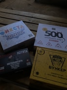 Four different board game boxes placed on a wooden surface. The designs feature colorful and graphic artwork. Each box appears to have distinct themes, represented by unique titles and illustrations.