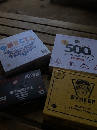 Four different board game boxes placed on a wooden surface. The designs feature colorful and graphic artwork. Each box appears to have distinct themes, represented by unique titles and illustrations.