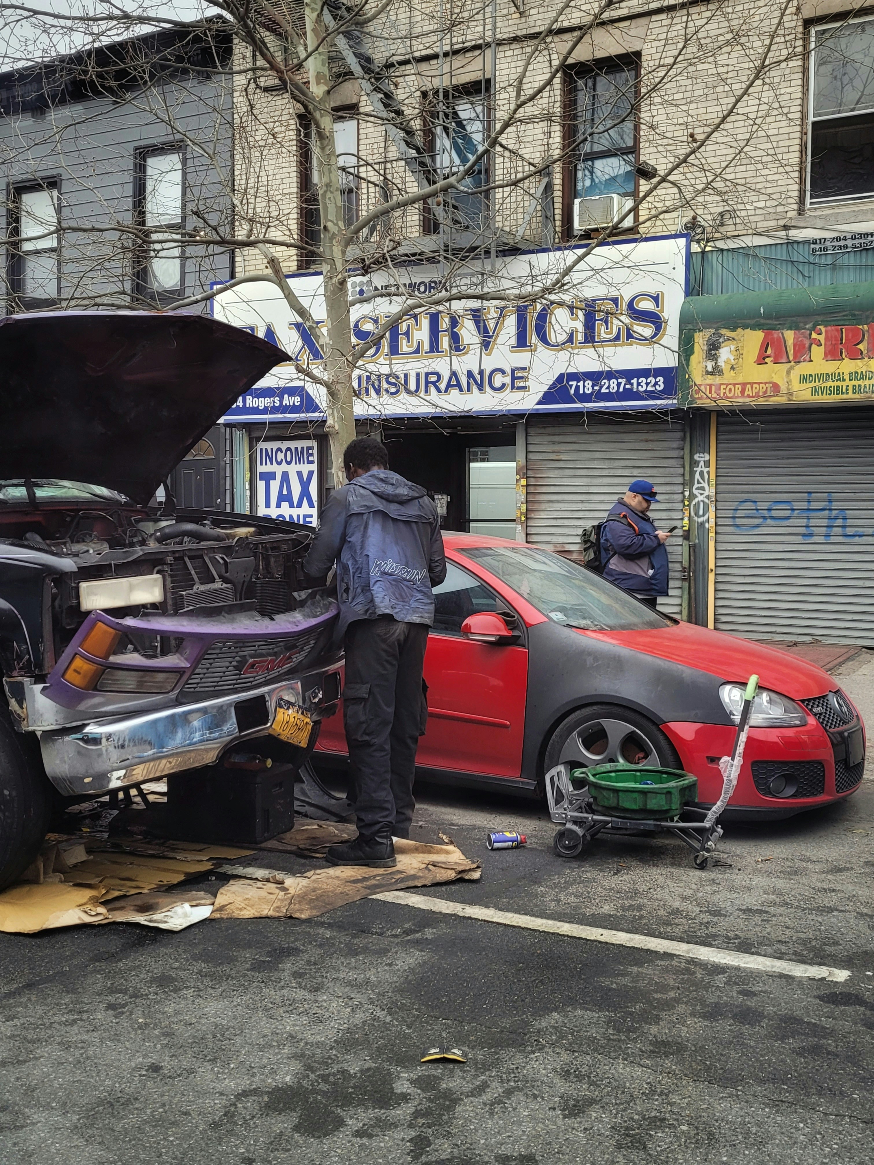 Two individuals are present near a vehicle with its hood open on a street in front of a building with various business signs. One person appears to be working on the engine, while the other is observing or engaged in some other task. A red car is parked beside them, and tools along with some automotive fluids are visible on the ground.