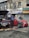 Two individuals are present near a vehicle with its hood open on a street in front of a building with various business signs. One person appears to be working on the engine, while the other is observing or engaged in some other task. A red car is parked beside them, and tools along with some automotive fluids are visible on the ground.