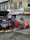 Two individuals are present near a vehicle with its hood open on a street in front of a building with various business signs. One person appears to be working on the engine, while the other is observing or engaged in some other task. A red car is parked beside them, and tools along with some automotive fluids are visible on the ground.