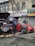 Two individuals are present near a vehicle with its hood open on a street in front of a building with various business signs. One person appears to be working on the engine, while the other is observing or engaged in some other task. A red car is parked beside them, and tools along with some automotive fluids are visible on the ground.