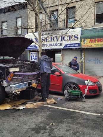 Two individuals are present near a vehicle with its hood open on a street in front of a building with various business signs. One person appears to be working on the engine, while the other is observing or engaged in some other task. A red car is parked beside them, and tools along with some automotive fluids are visible on the ground.
