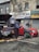 Two individuals are present near a vehicle with its hood open on a street in front of a building with various business signs. One person appears to be working on the engine, while the other is observing or engaged in some other task. A red car is parked beside them, and tools along with some automotive fluids are visible on the ground.