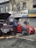 Two individuals are present near a vehicle with its hood open on a street in front of a building with various business signs. One person appears to be working on the engine, while the other is observing or engaged in some other task. A red car is parked beside them, and tools along with some automotive fluids are visible on the ground.