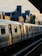 A metropolitan train travels along elevated tracks, partially illuminated by the warm light of a setting or rising sun. The surrounding environment includes urban buildings with a mix of modern and industrial architectural styles, casting long shadows.