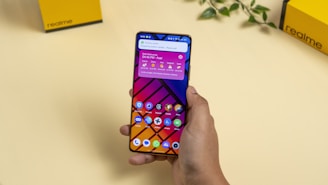 A hand holding a smartphone displaying a colorful home screen with various app icons against a geometric background. The screen shows a time, date, weather widget, and notifications. In the background, there are yellow boxes with 'realme' branding and a small plant on a beige surface.