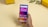 A hand holding a smartphone displaying a colorful home screen with various app icons against a geometric background. The screen shows a time, date, weather widget, and notifications. In the background, there are yellow boxes with 'realme' branding and a small plant on a beige surface.