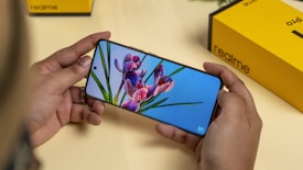 A person holds a smartphone displaying an image of flowers with a vivid screen resolution. A yellow and black Realme product box is visible in the background on the table.